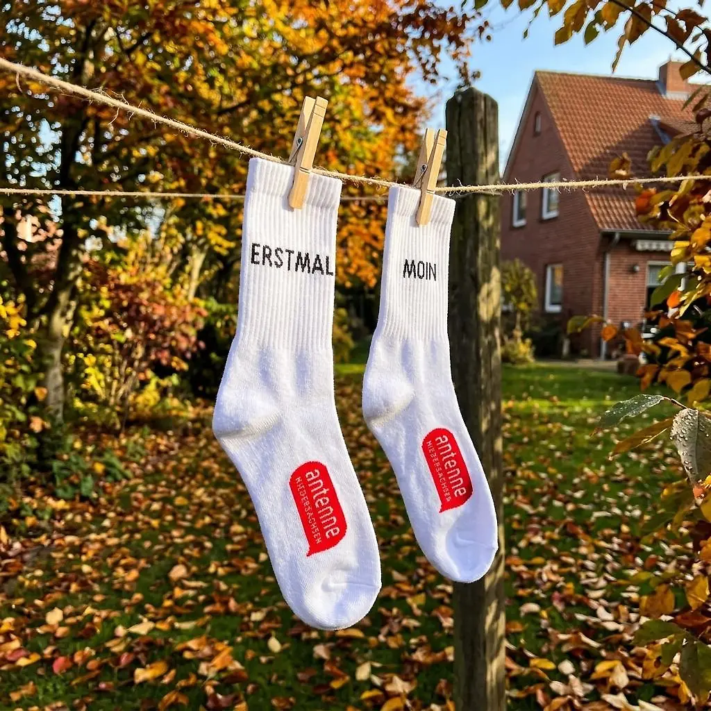 Weiße Socken mit der Aufschrift "ERSTMAL MOIN" und einem roten Logo von Antenne Niedersachsen hängen von einem herbstlichen Baum. Im Hintergrund ist ein rotes Backsteinhaus