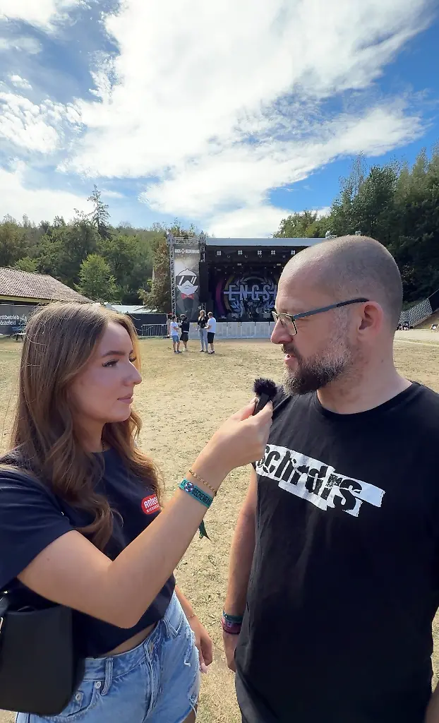 Eine Frau interviewt einen Mann mit einem Mikrofon vor einer Bühne im Freien.