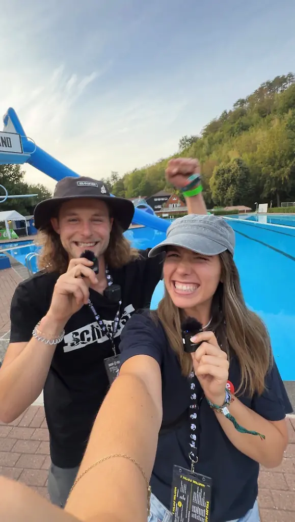 Zwei Personen lächeln und posieren mit Mikrofonen vor einem leeren Pool.