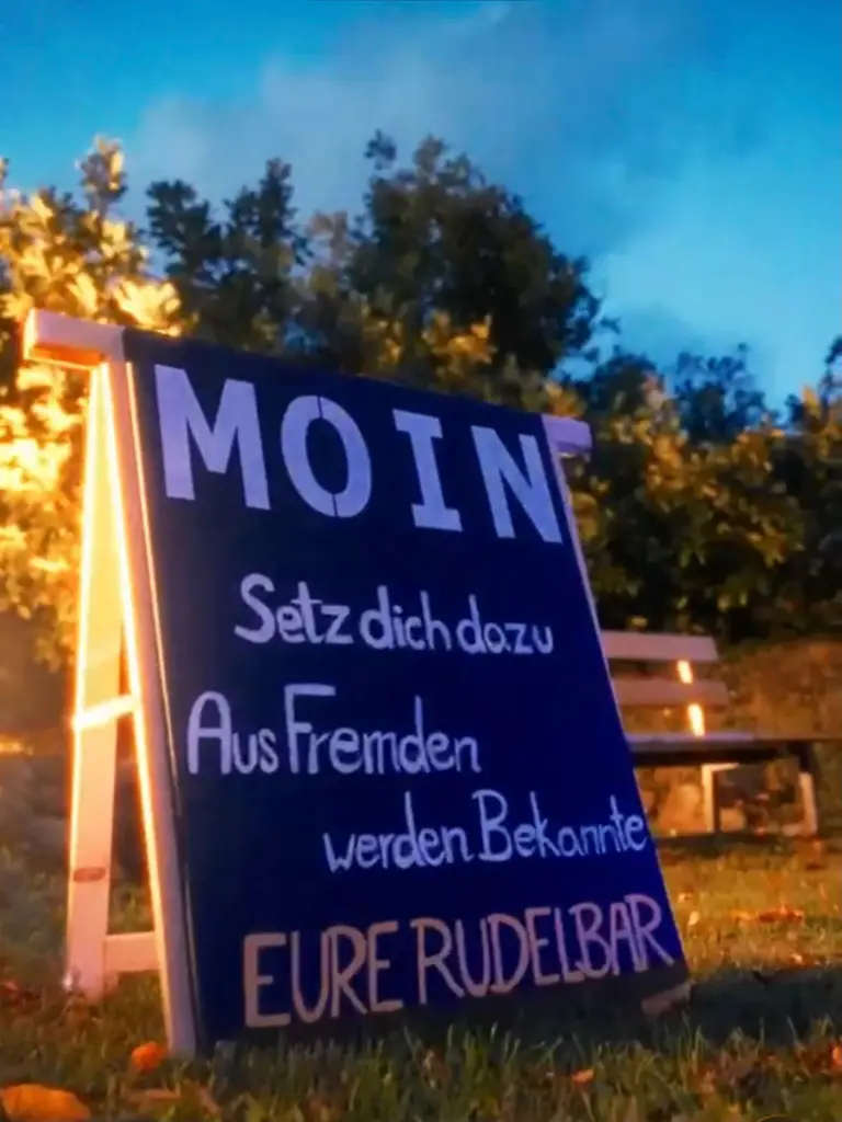 Ein Schild mit der Aufschrift "MOIN Setz dich dazu Aus Fremden werden Bekannte EURE RUDELBAR" steht auf einer Wiese.