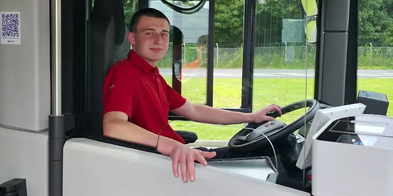 Ein junger Mann sitzt in einem Bus am Fahrersitz und schaut in die Kamera.