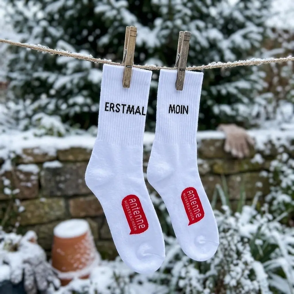 Weiße Socken mit der Aufschrift "ERSTMAL MOIN" und einem roten Logo von Antenne Niedersachsen hängen von einer schneebedeckten Mauer auf einer Leine